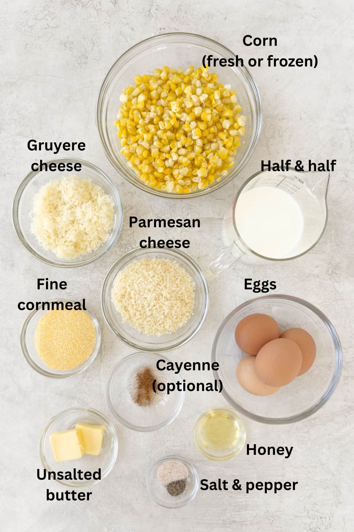 Ingredients for corn souffle in glass prep bowls on a counter with corn kernels, cheeses, eggs, cornmeal. 
