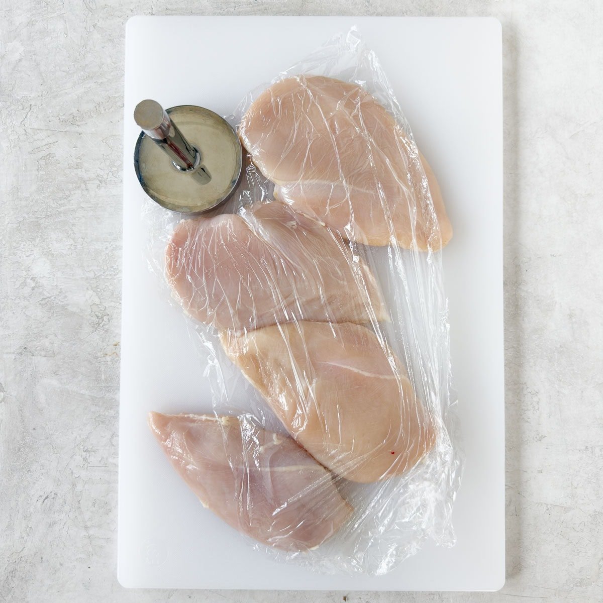 Pounding boneless skinless chicken breasts on a cutting board covered in plastic.