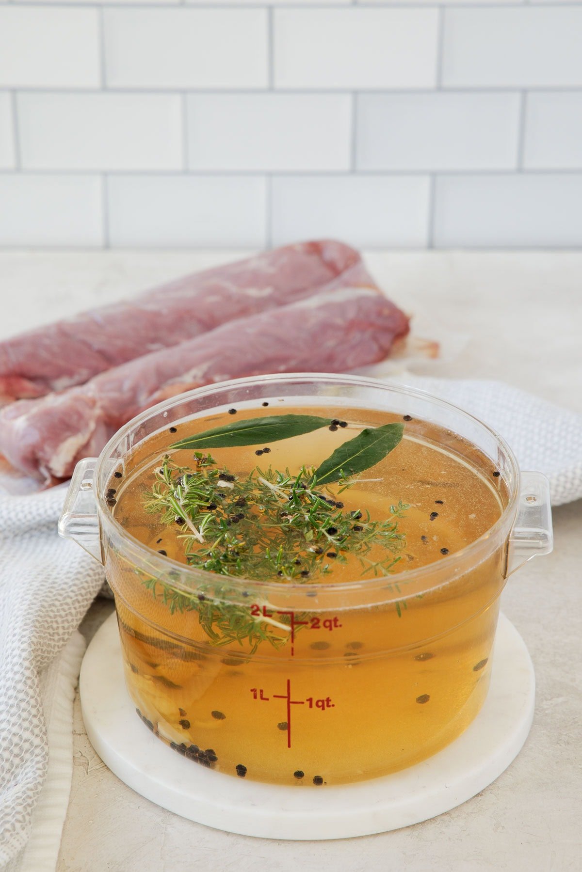 A container of clear golden pork brine with fresh herbs and crushed garlic.