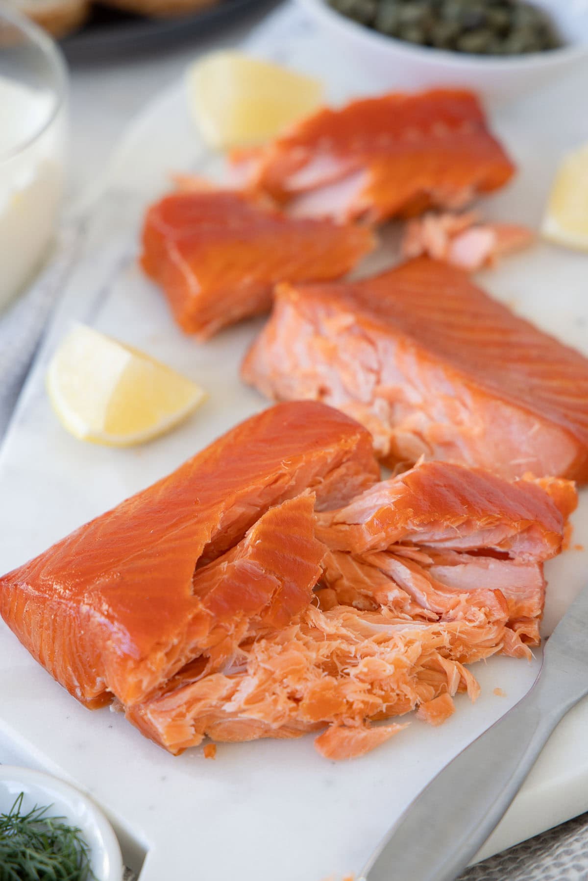 Bright orange smoked salmon chunks on a white board with lemon wedges and capers in the back.