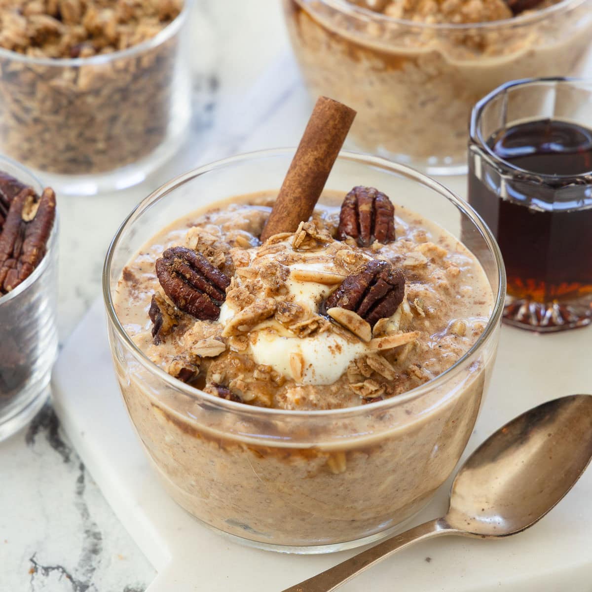 Pumpkin spice overnight oats in a glass serving bowl with cinnamon, pecans, maple syrup, and spoon.