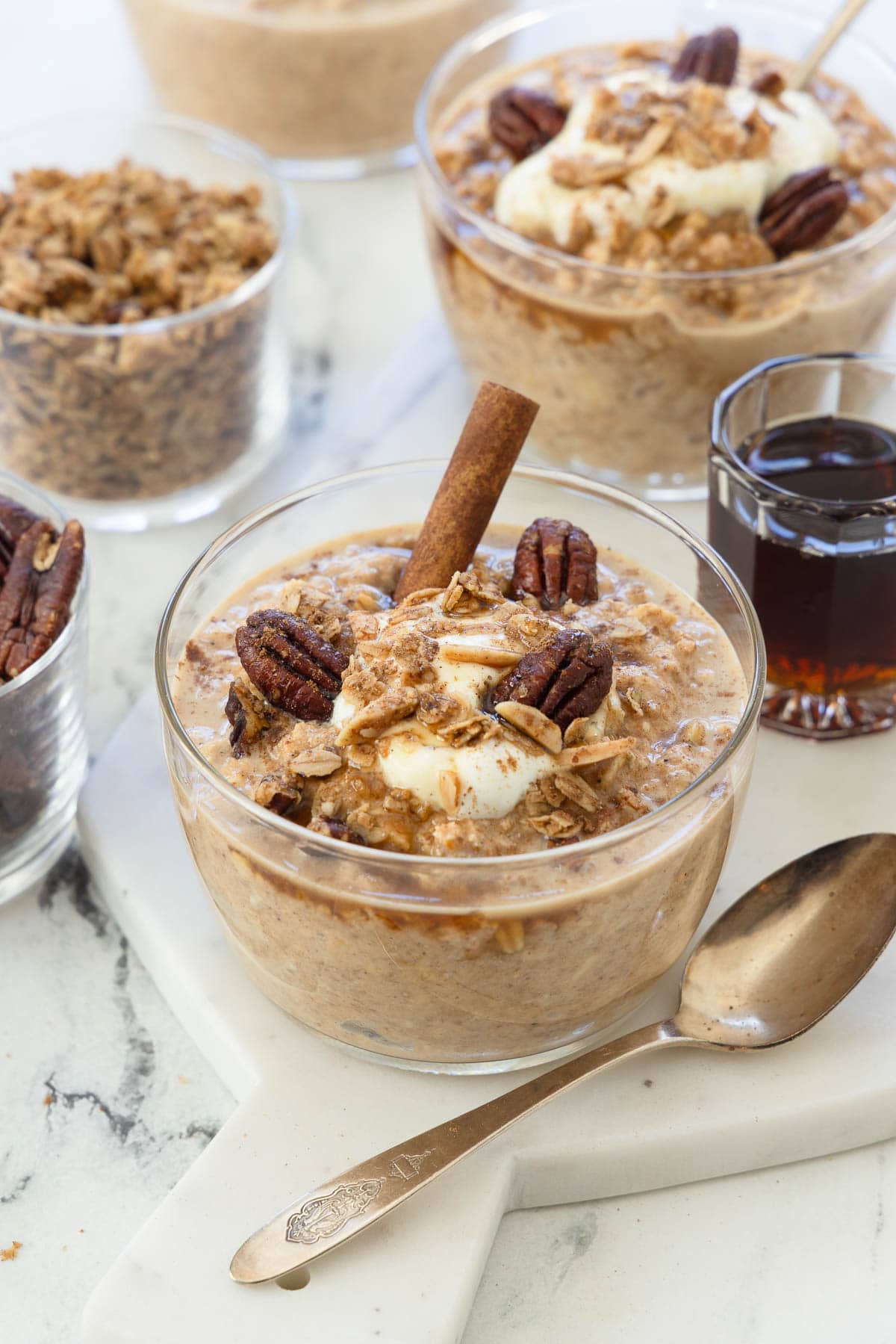 Bowls of pumpkin overnight ots with cinnamon stick, pecans, yogurt, and a silver spoon.
