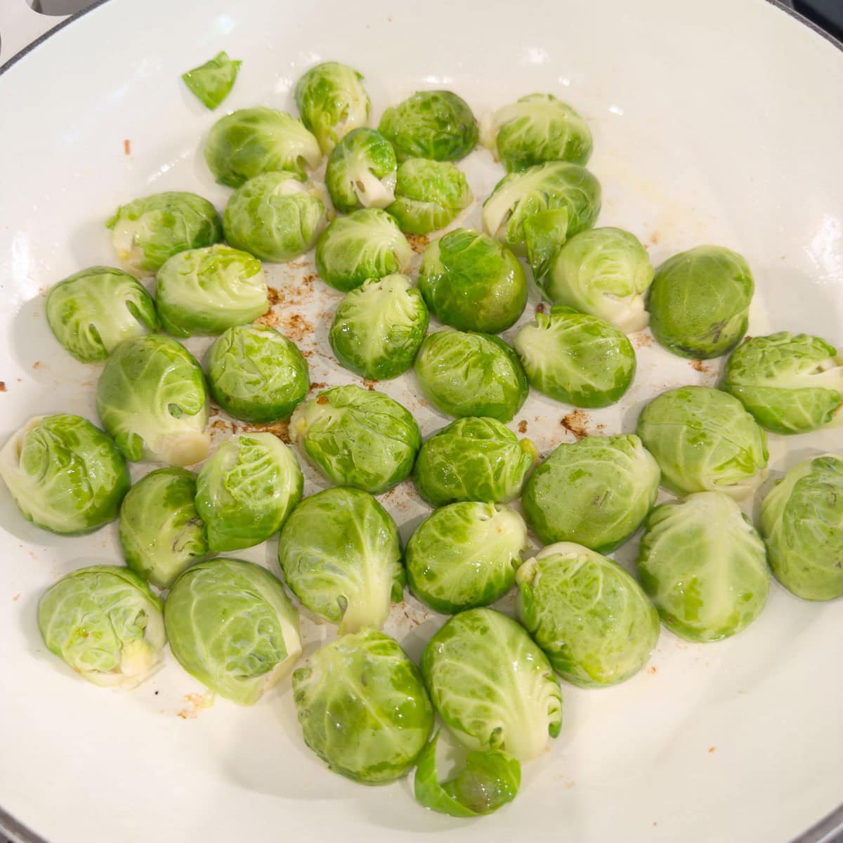 Halved Brussels sprouts browning in a white pan on sthe stovetop.