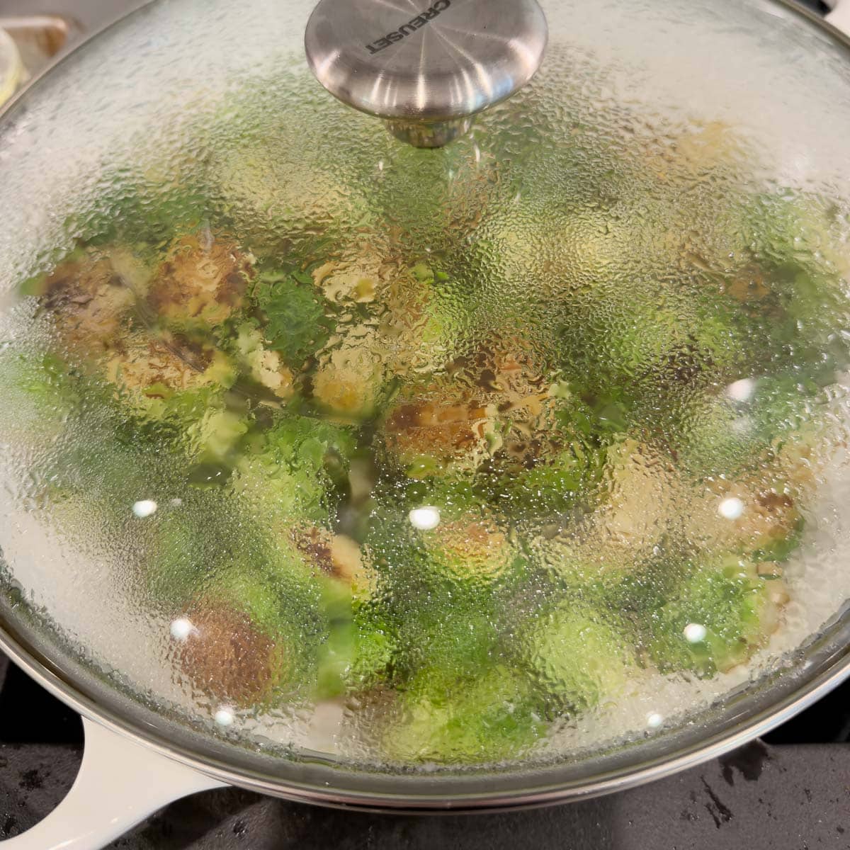A wide shallow pan with a glass lid, with green vegetables inside on stovetop.