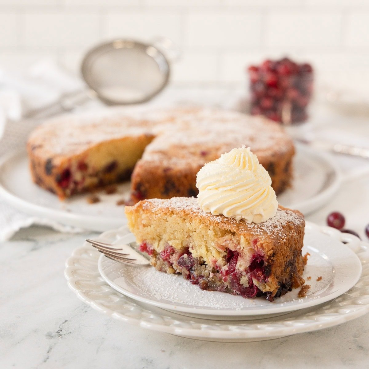 A slice of nantucket cranberry pie on a plate topped with whipped cream, whole cake behind.