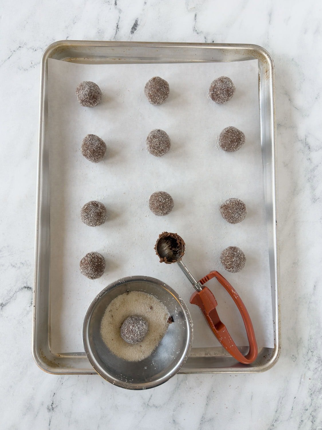 Chocolate cookie dough balls on a parchment lined baking sheet with a cookies scoop.