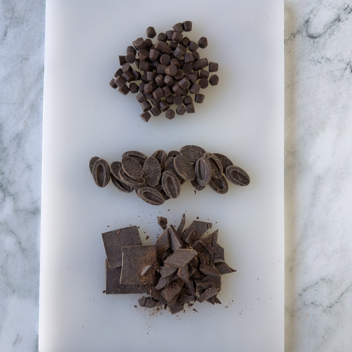 Forms of dark chocolate on a white cutting board.
