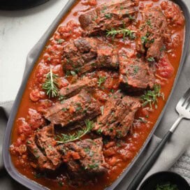 Rich red sauce with pot roast nestled into the vegetables and herbs on a gray plate.