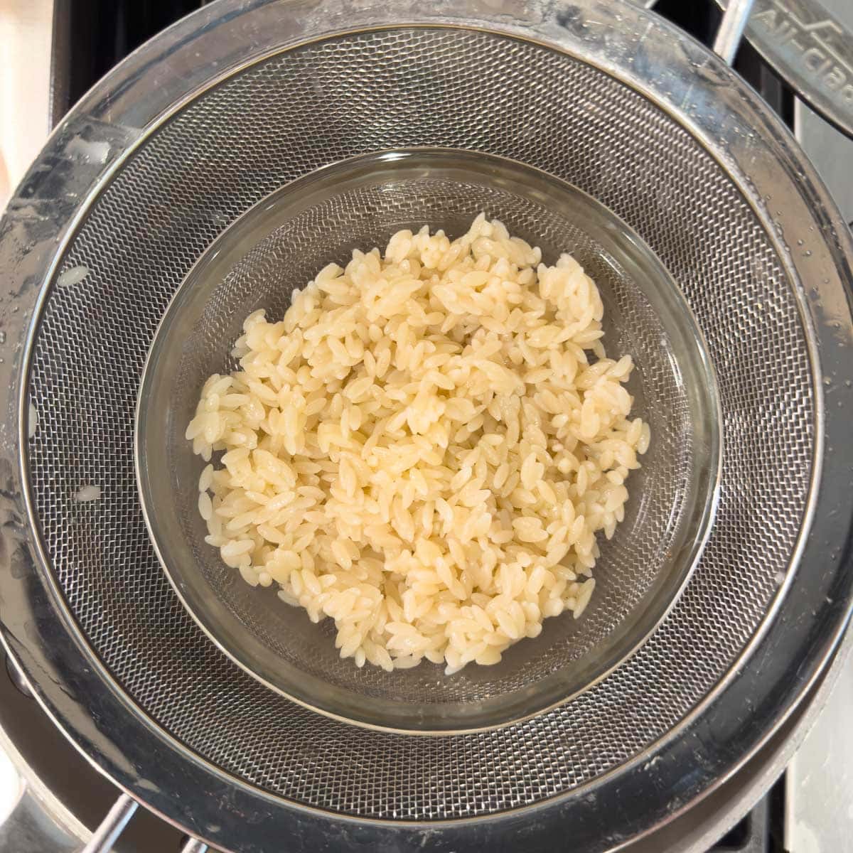 Cooked orzo pasta in a sieve over a bowl being rinsed and drained.