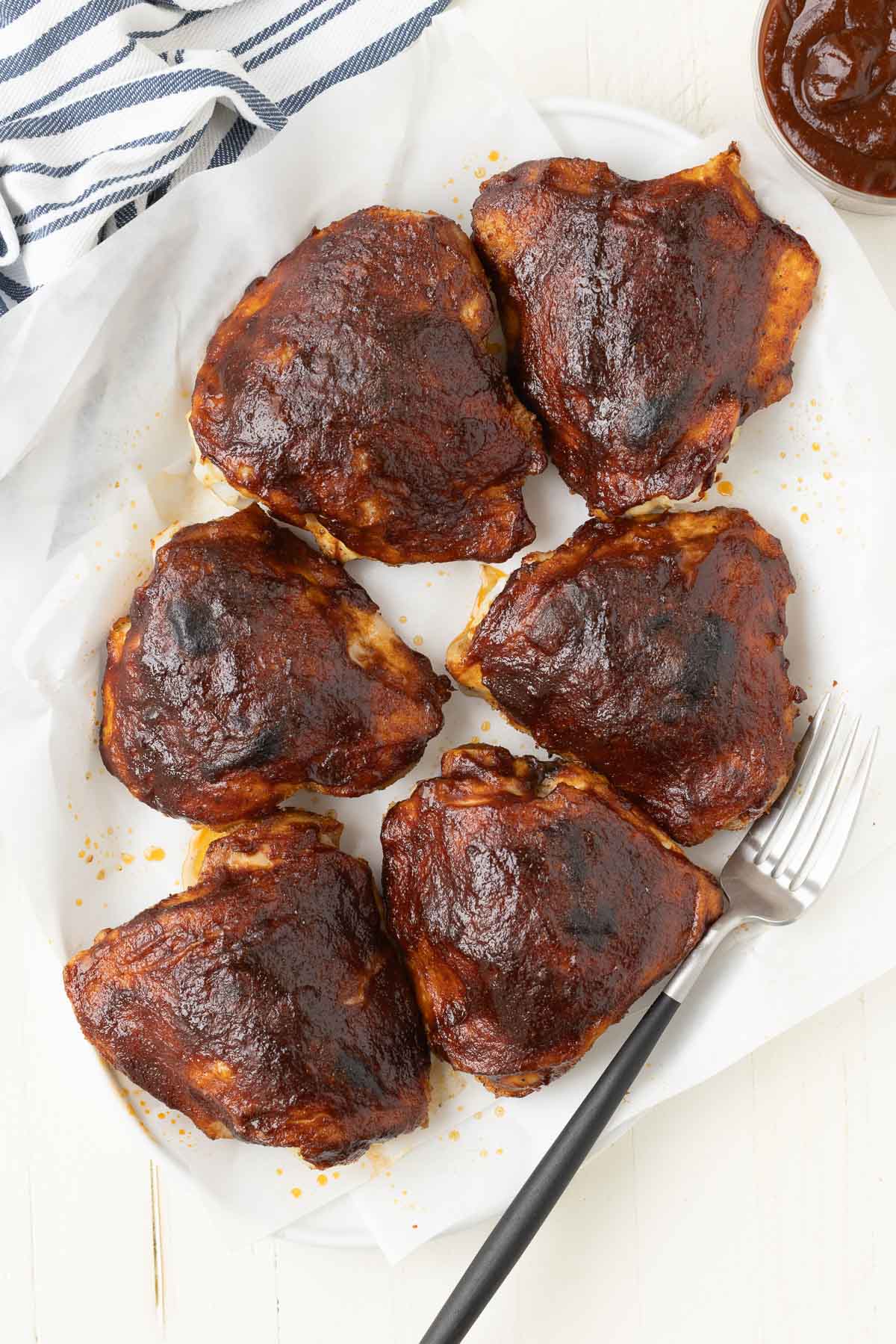 An oval platter with crinkled parchment and 6 glossy BBQ chicken thighs with serving fork.