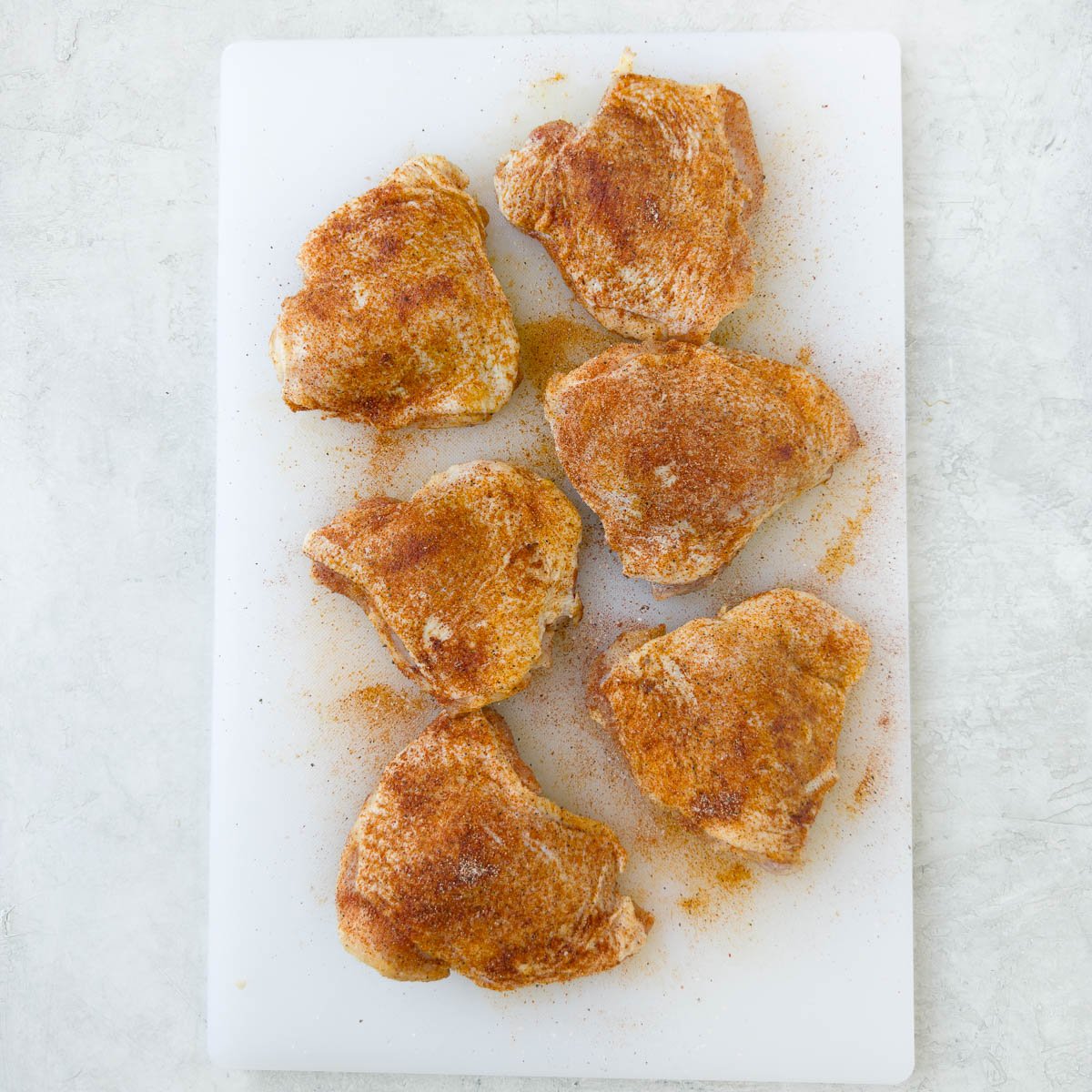 Six seasoned chicken thighs on a white cutting board.