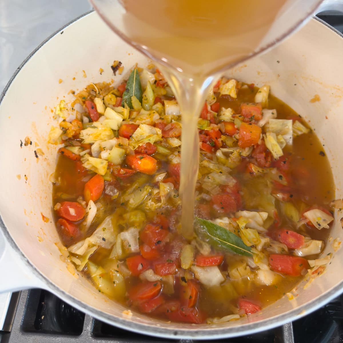 Golden chicken broth pouring into a pot of vegetables for soup.