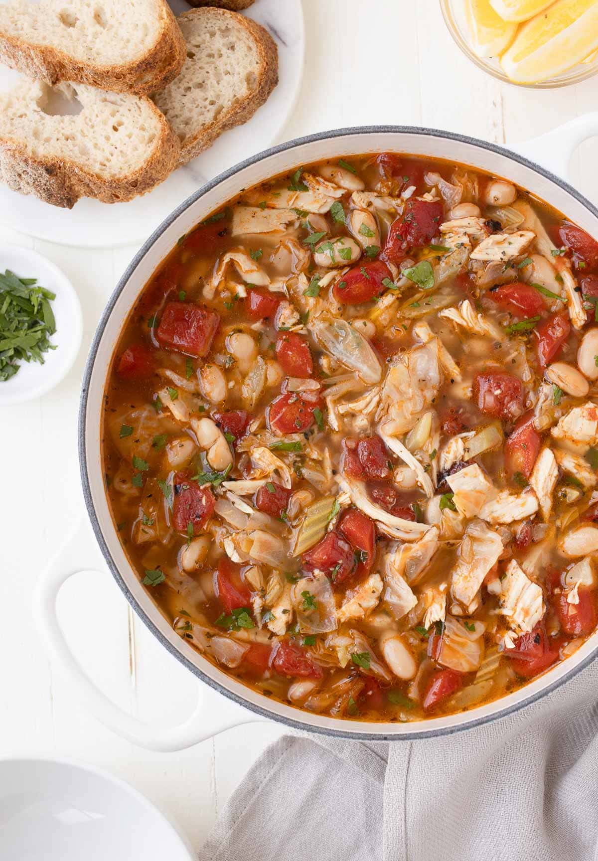A pot of colorful cabbage and chicken soup with tomatoes, carrots, beans, and herbs, with sliced bread.