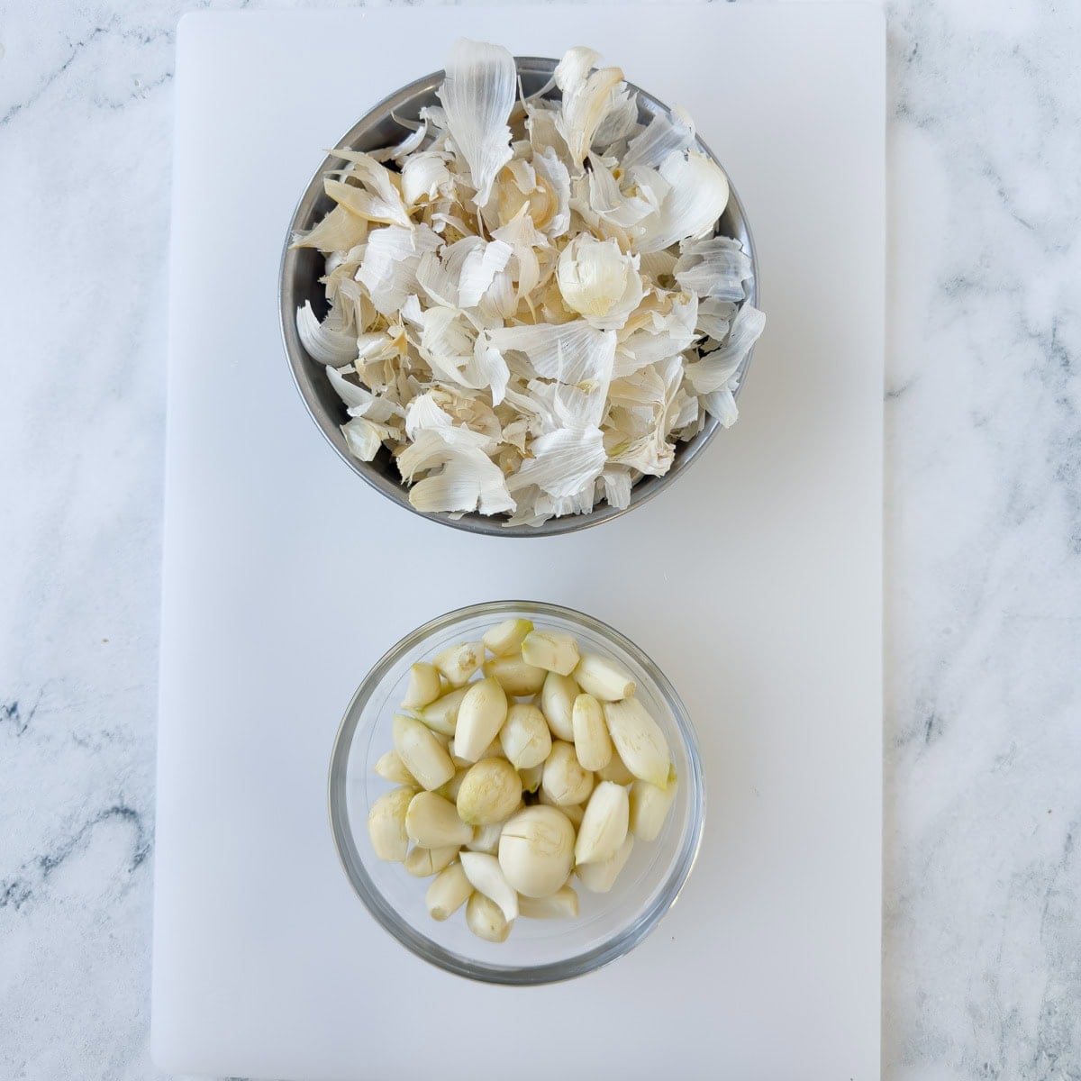 A glass bowl of whole peeled garlic cloves and a bowls of papery peels.
