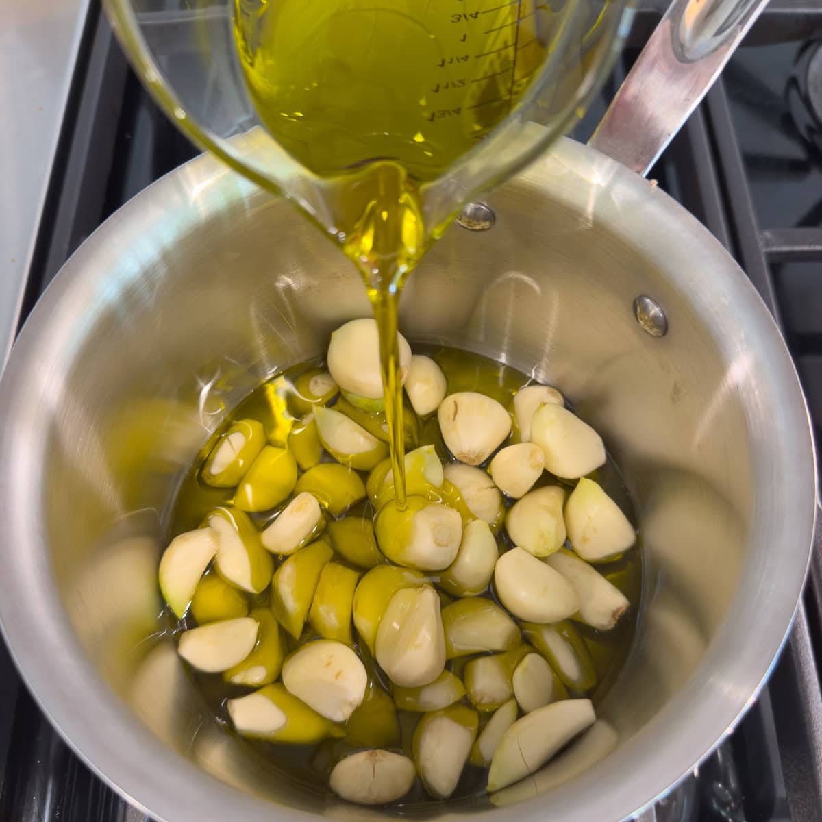 Pouring golden olive oil into a small pan over whole garlic cloves.