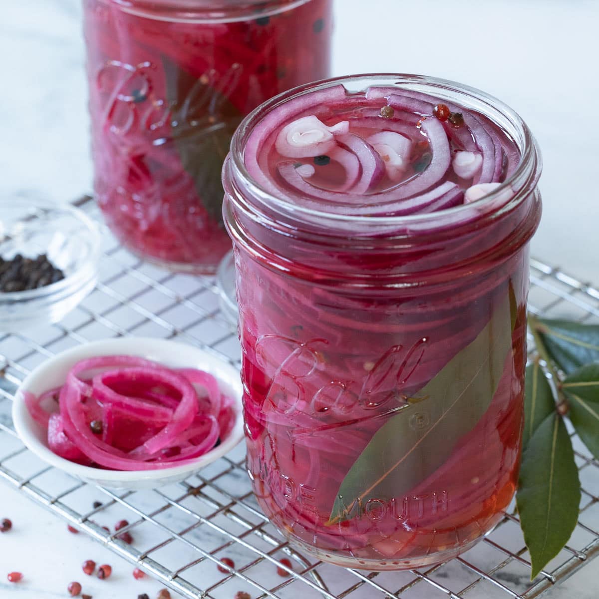 A jar of bright pink pickled onions with a small bowl of them beside, and bay leaves.