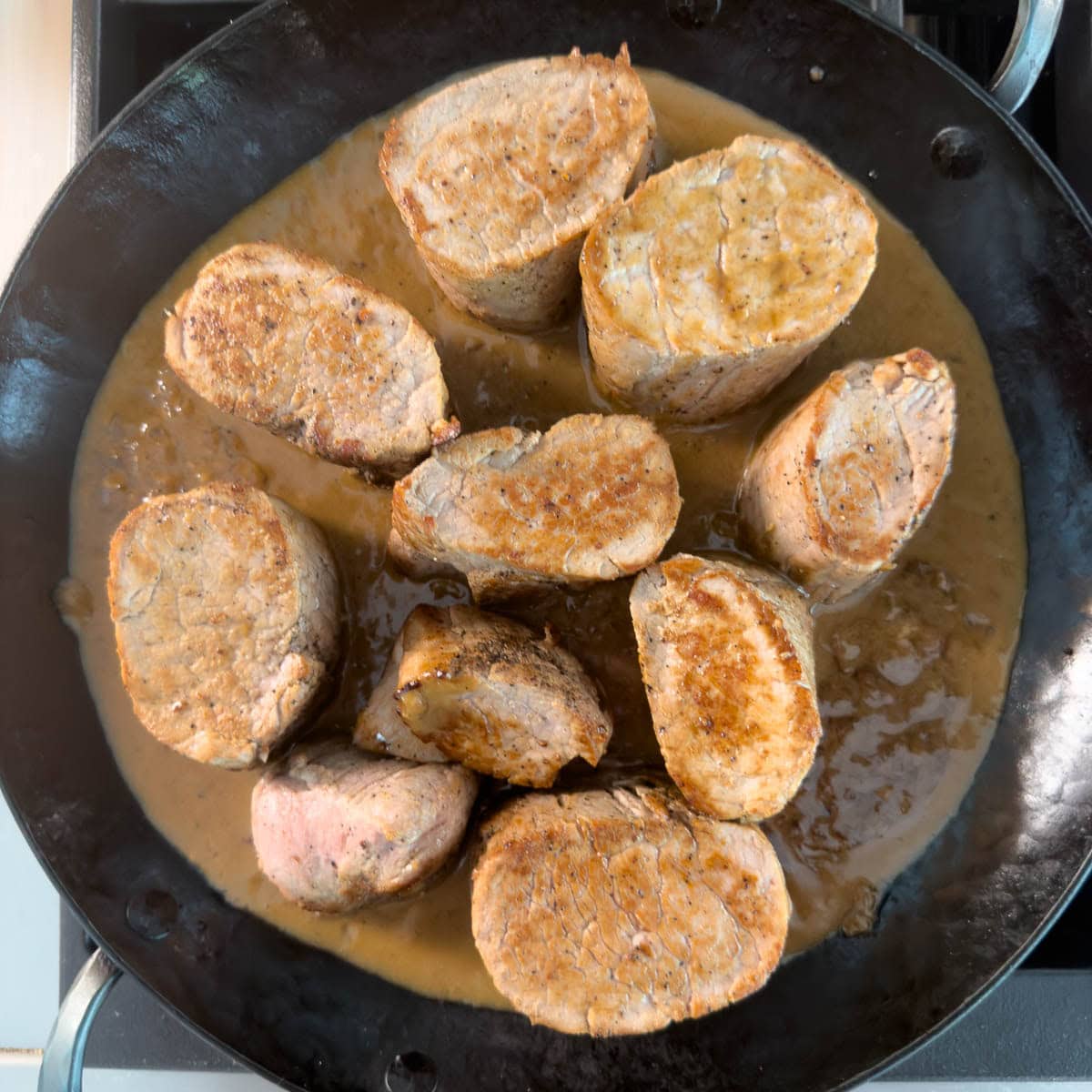 Finished pork tenderloin medallions in a golden brown creamy pan sauce in a black pan.