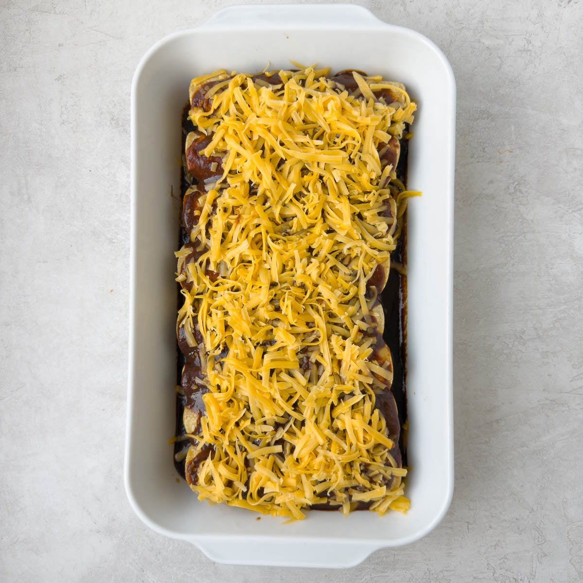 Grated cheddar cheese covering the top of beef enchiladas in a casserole, ready to bake.