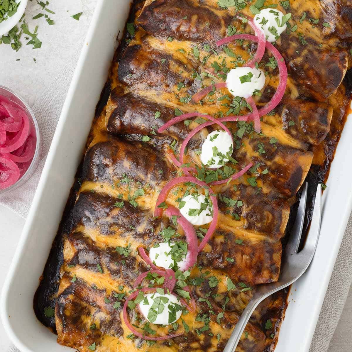 A baking dish full of shredded beef enchiladas with a deep red enchilada sauce cilantro, and pickled onions.