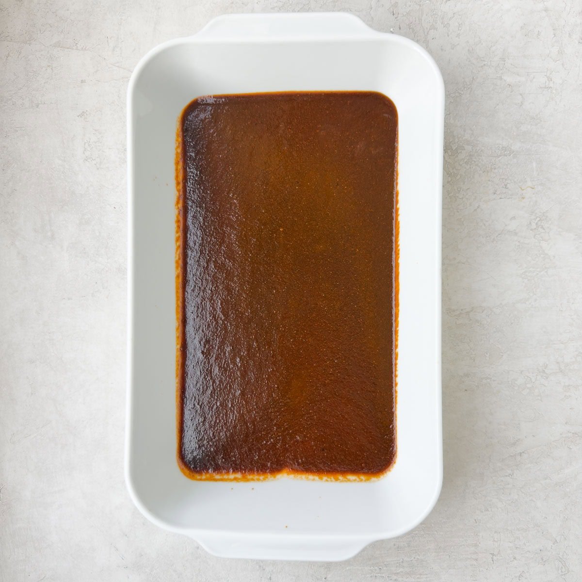 A white rectangular casserole dish with deep red enchilada sauce on the bottom.
