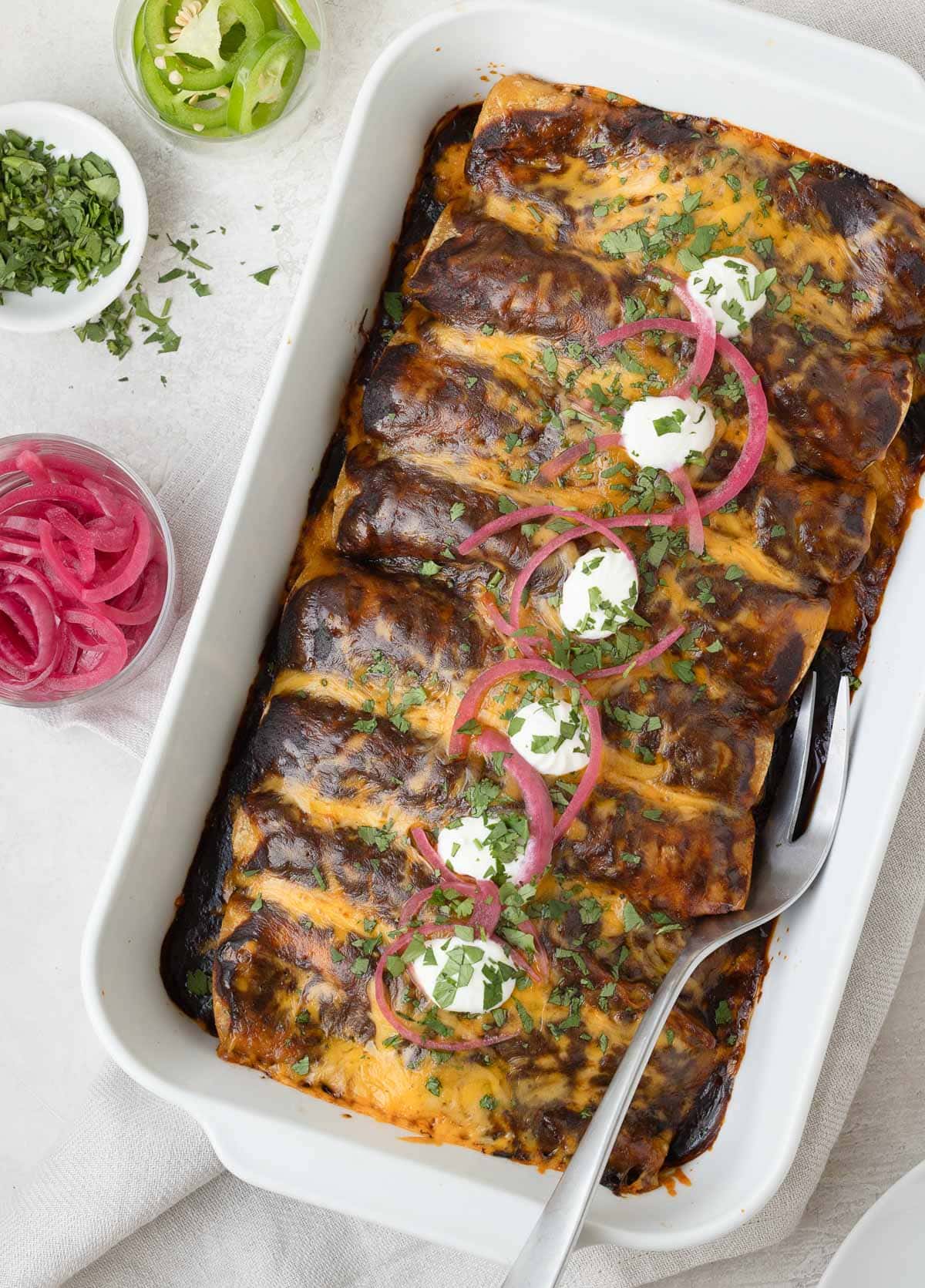 A casserole dish of shredded beef enchiladas with a deep dark red chili sauce, pink pickled onions, and cilantro.