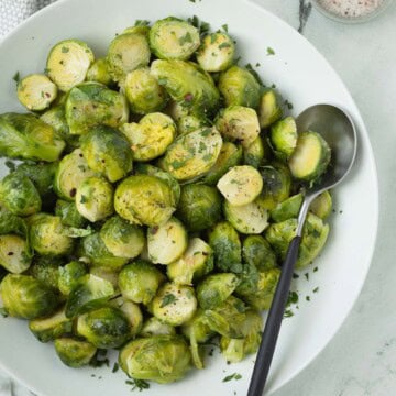 A white bowl of brght green Brussels sprouts tossed with olive oil, salt, pepper and red pepper flakes.