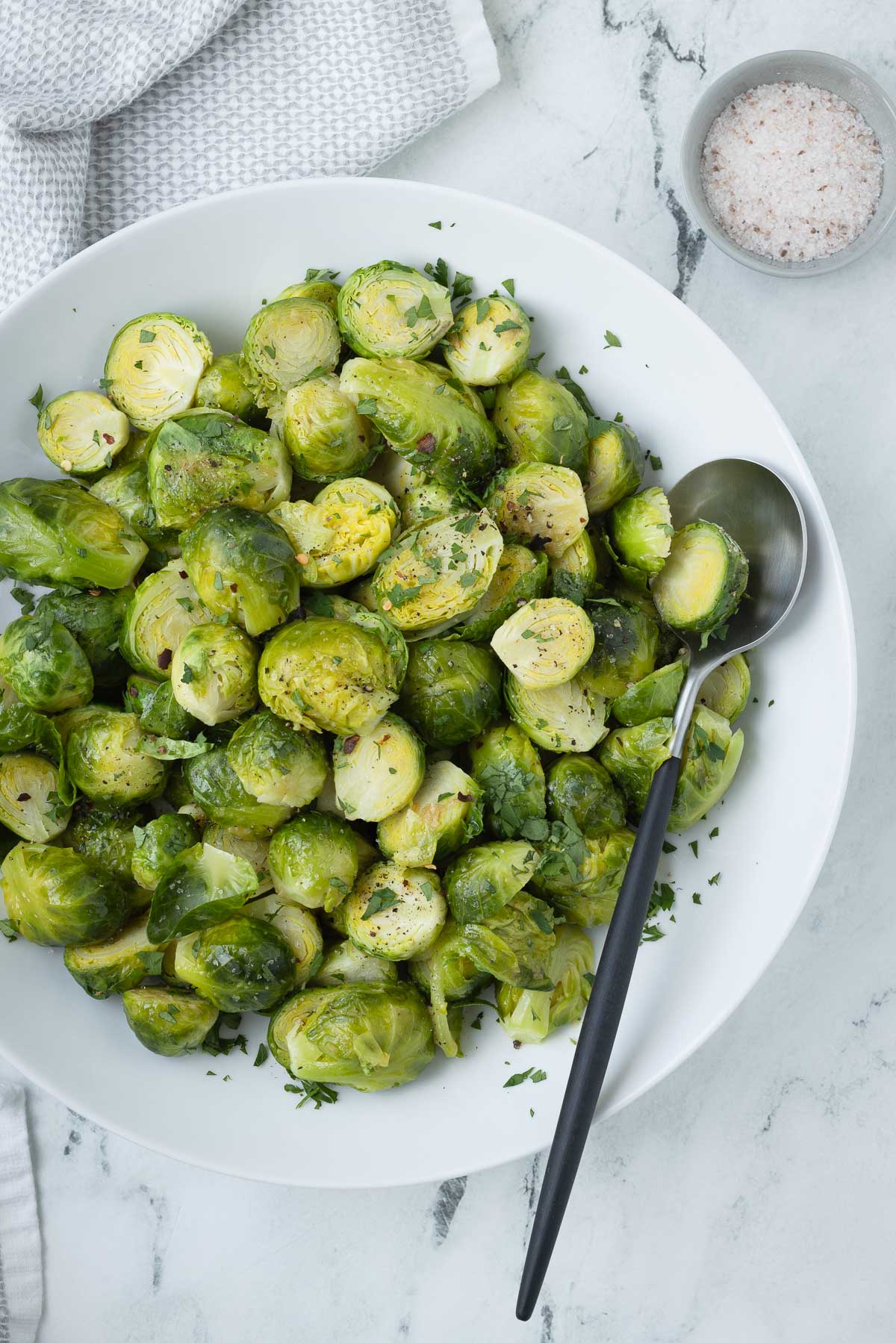 A white round bowl of bright green steamed Brussels sprouts with a serving spoon. 