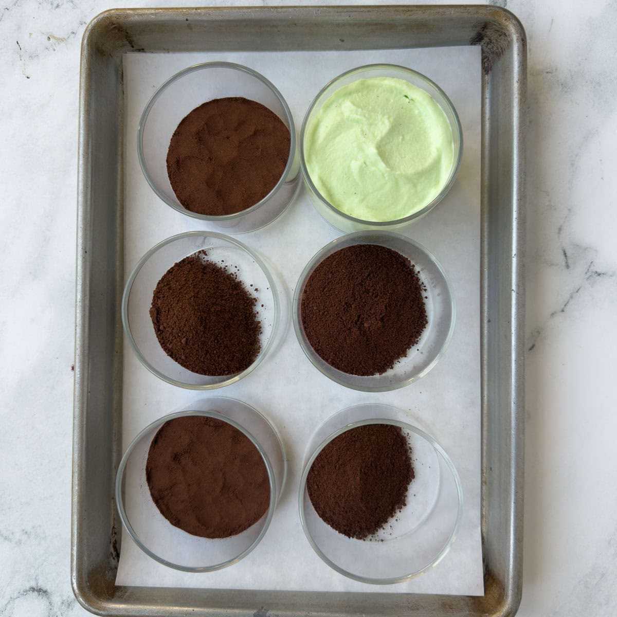 Six small glass bowls being filled with chocolate cookie crumbs and grasshopper mousse.