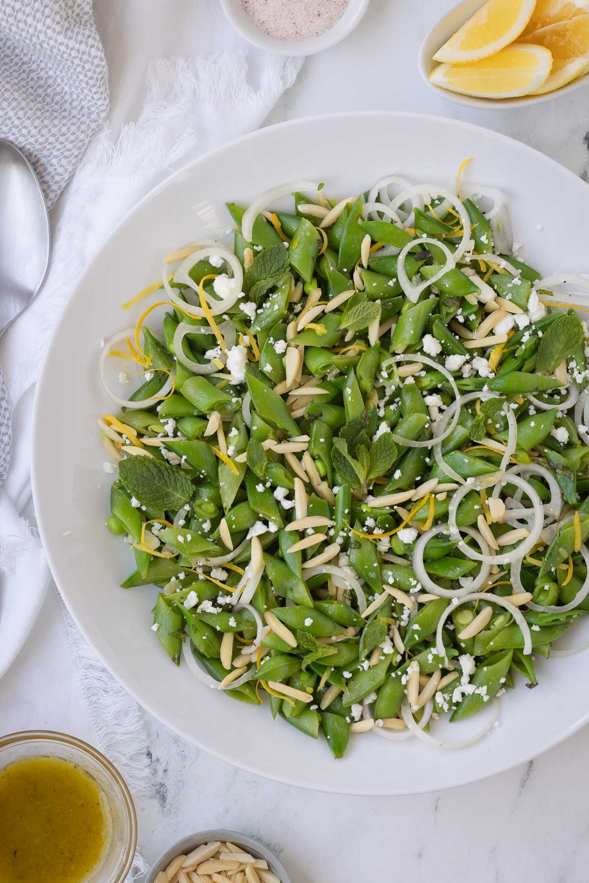 A white bowl of bright green sugar snap pea salad with almonds, lemon, feta, and mint. 