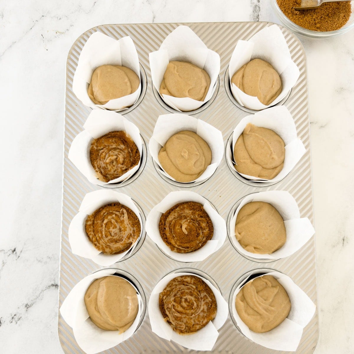 Cinnamon muffin batter portioned into tulip papers in a muffin pan.