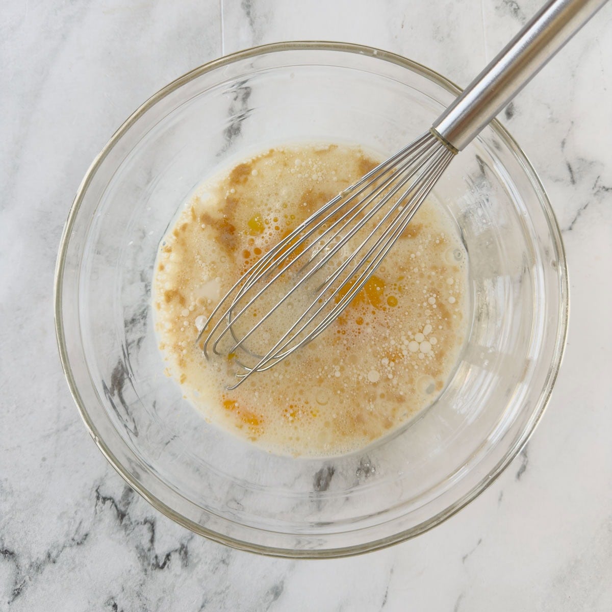 Mixing eggs, milk, oil, and wet ingredients for muffins in a glass bowl with a whisk.