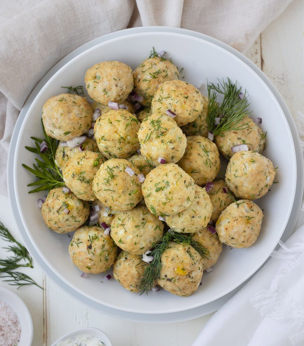 A white bowl of golden Greek chicken meatballs sprinkled with red onion and herbs.