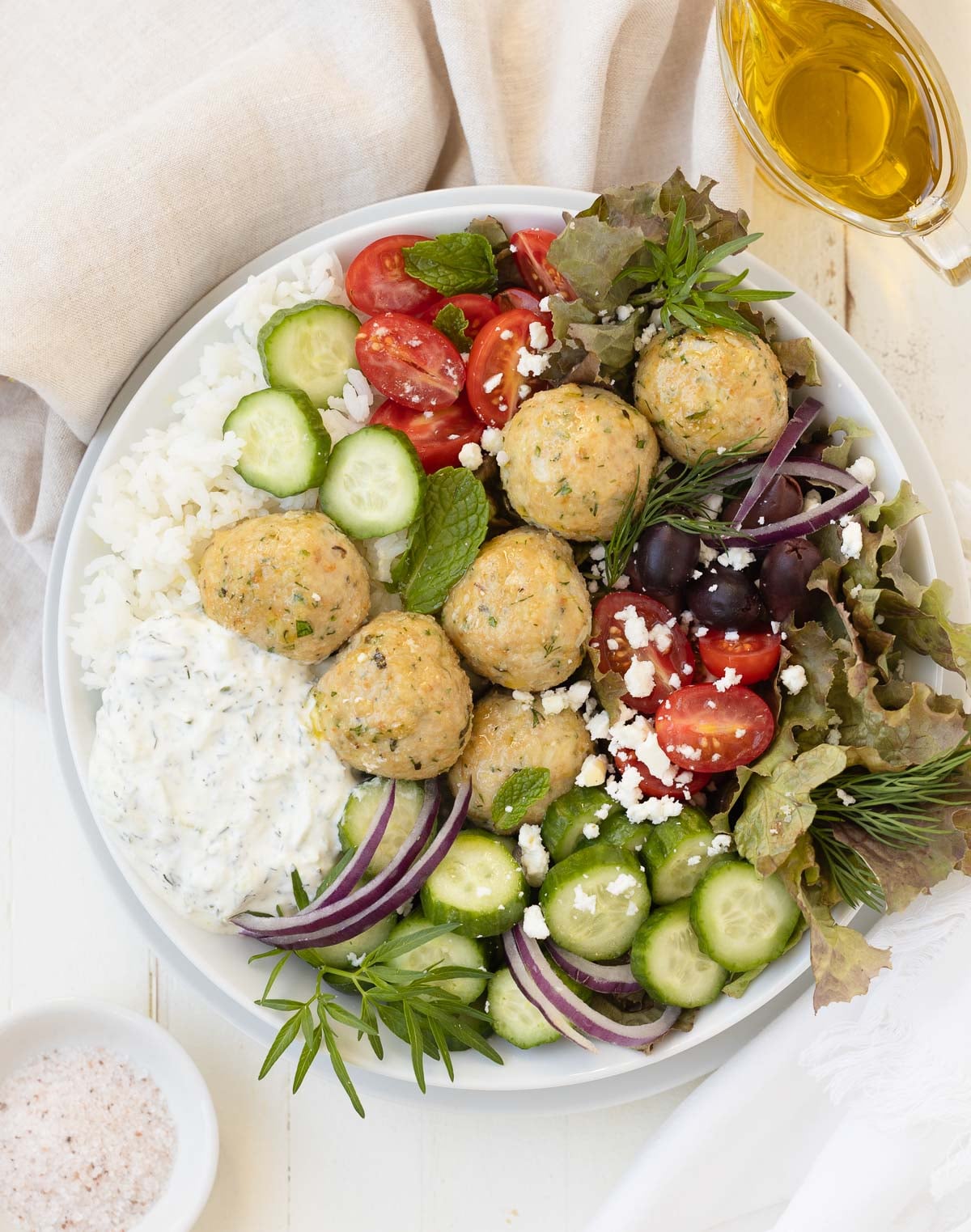 A grain bowl filled with greek chicken meatballs, rice, tomatoes, cucumbers, olive, and tzatziki sauce.