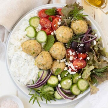 A rice bowl of Greek chicken meatballs with cucumbers, tomatoes, tzatziki, herbs, and olives with olive oil.