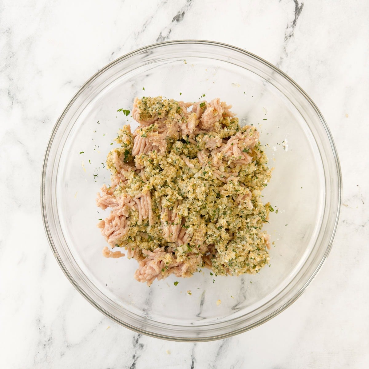 Adding pink chicken meat to a bowl of breadcrumbs, herbs, egg, and more as a chicken meatballs mix.
