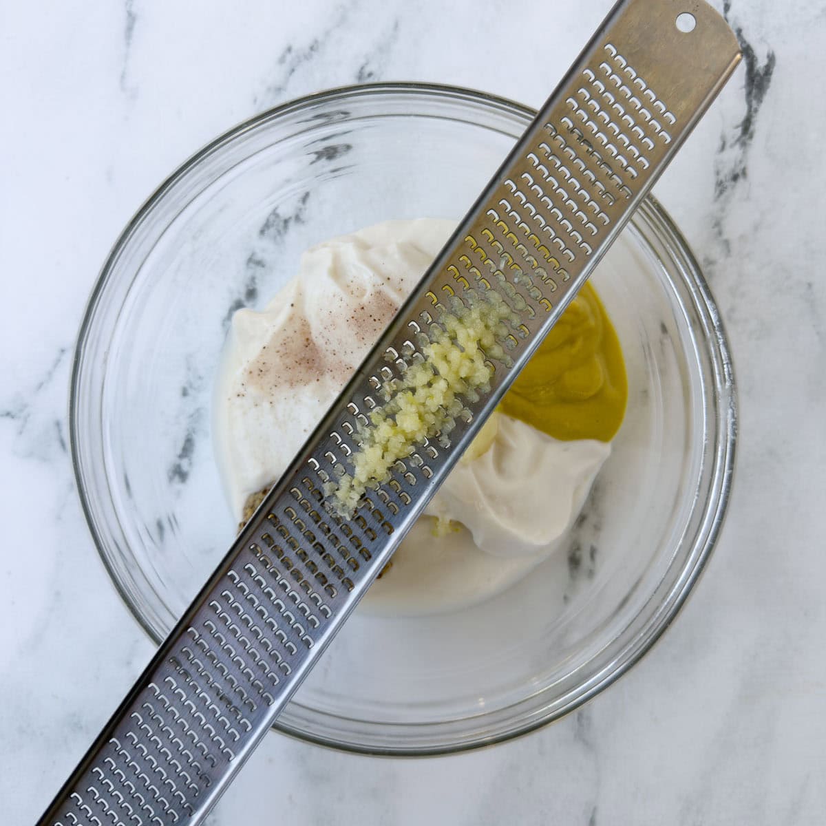 A microplane zester with garlic paste over a glass bowl.