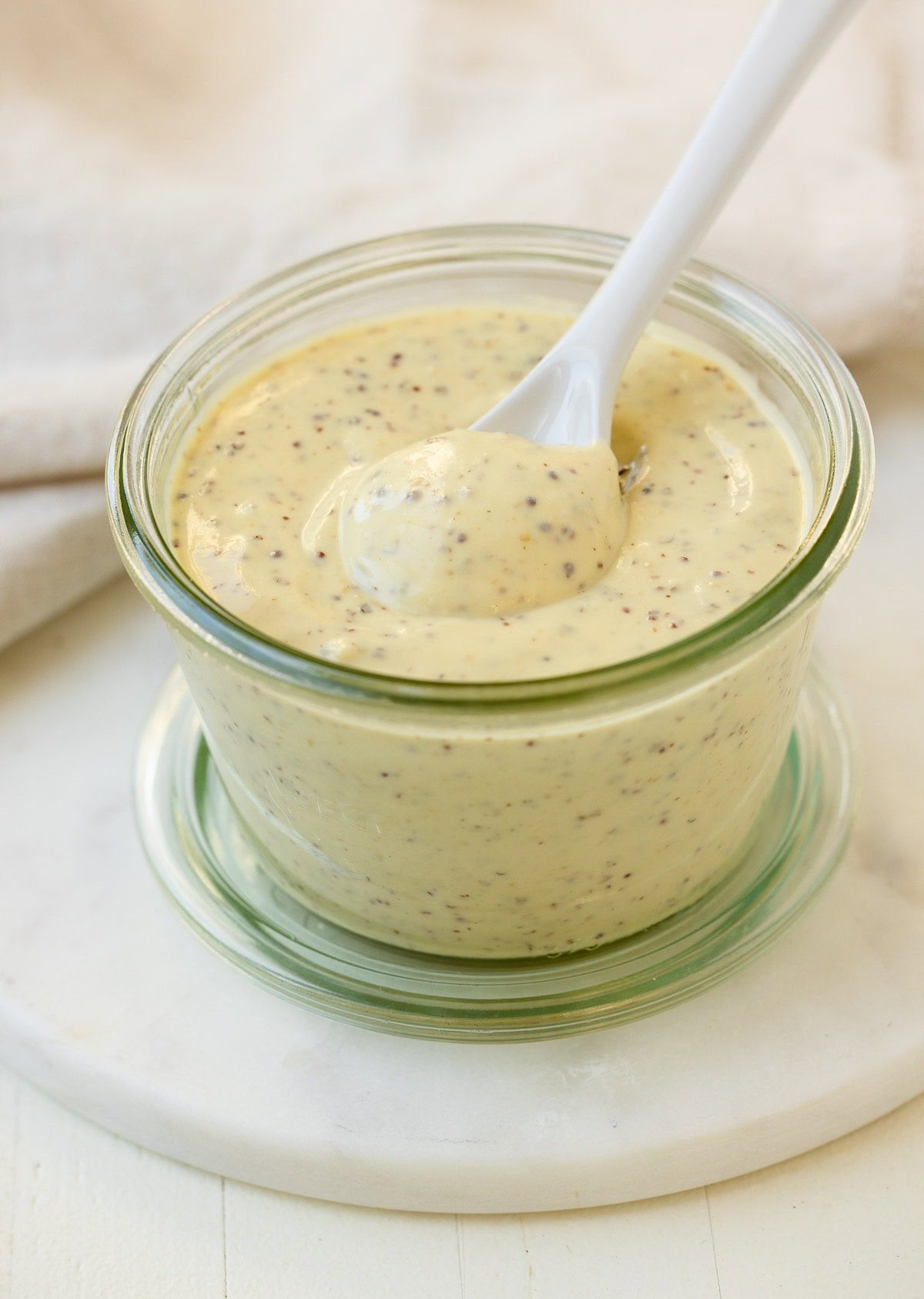 A jar of cream Dijon mustard aioli sauce with a white ceramic spoon. 