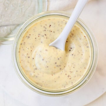 Pale yellow Dijon mustard aioli sauce in a glass jar with a white spoon.