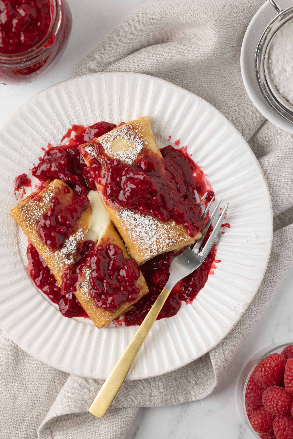 A plate of blintzes with cheese and ruby raspberry sauce with a gold fork.