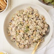 A serving bowl of chickpea tuna salad with a serving spoon, nearby lemon wedges, crackers, and greens.
