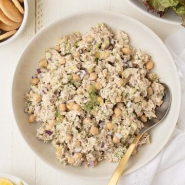 A serving bowl of chickpea tuna salad with a serving spoon, nearby lemon wedges, crackers, and greens.