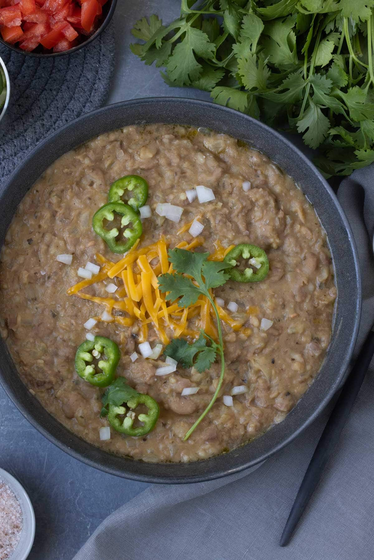 A bowl of refried-style pinto beans topped with grated cheese and jalapeno slices, cilantro sprigs.