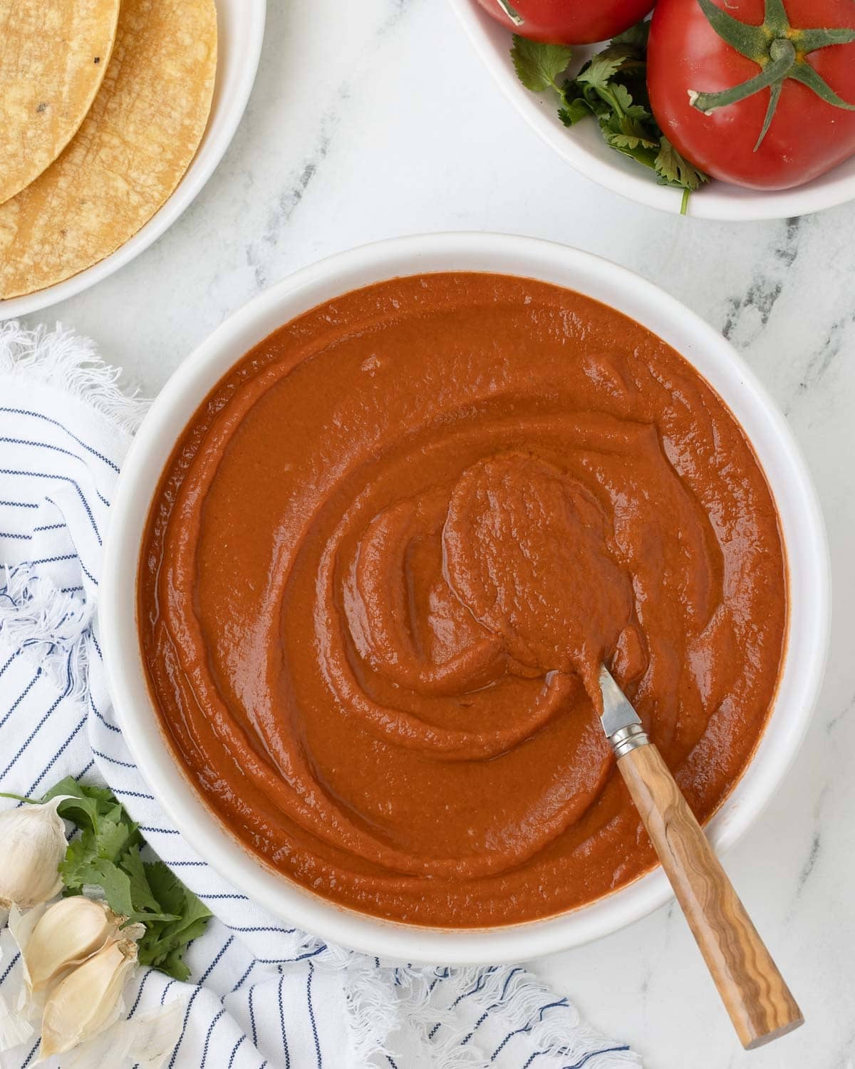 A bowl of deep red ranchero sauce in a white bowl with spoon, garlic cloves, and tortillas.