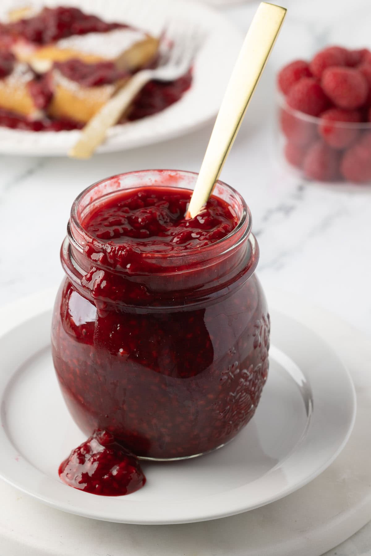 A jar of chunky raspberry sauce with a spoon on a white plate, sauce dripping down side.