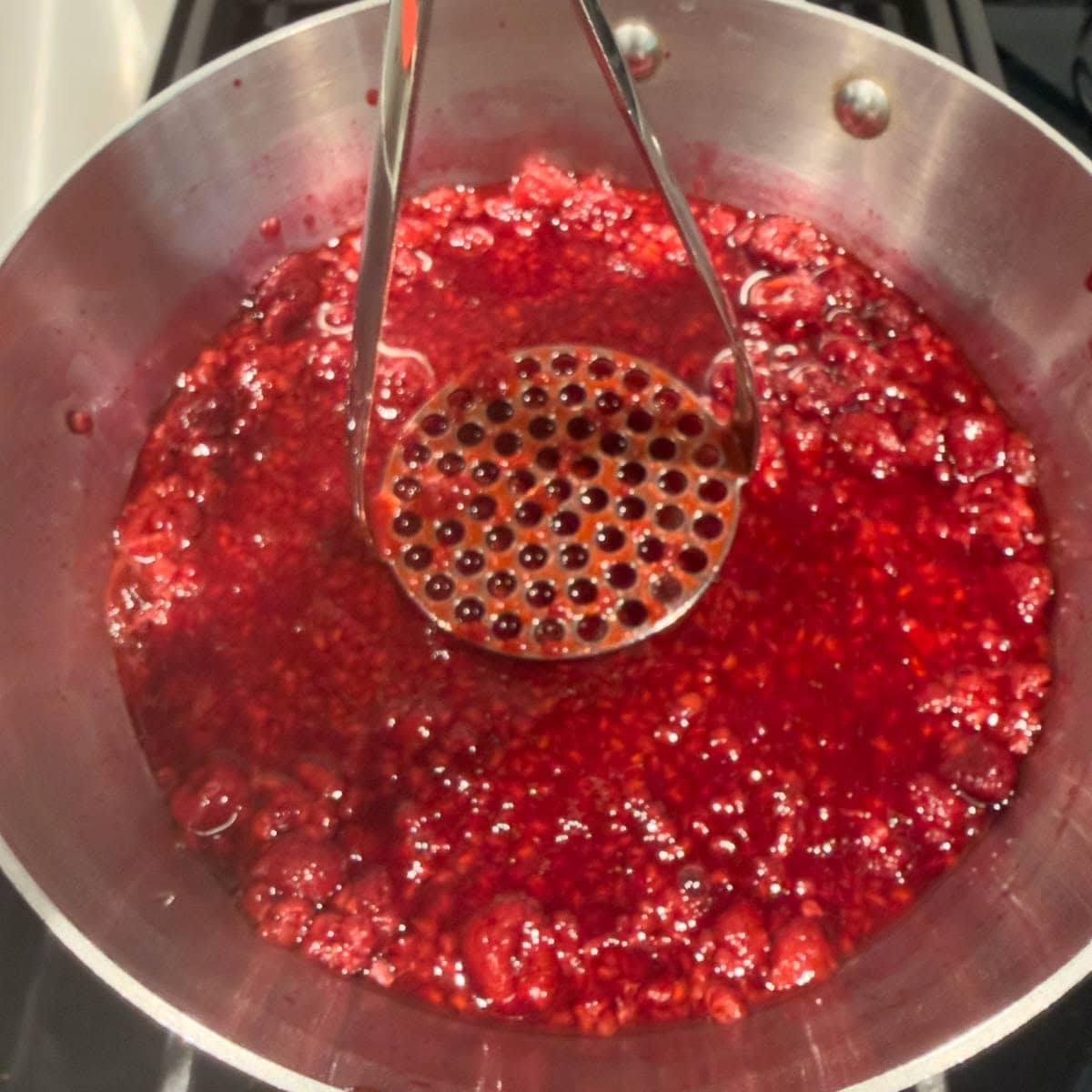 Mashing cooking raspberries with a potato masher to break up the berries.