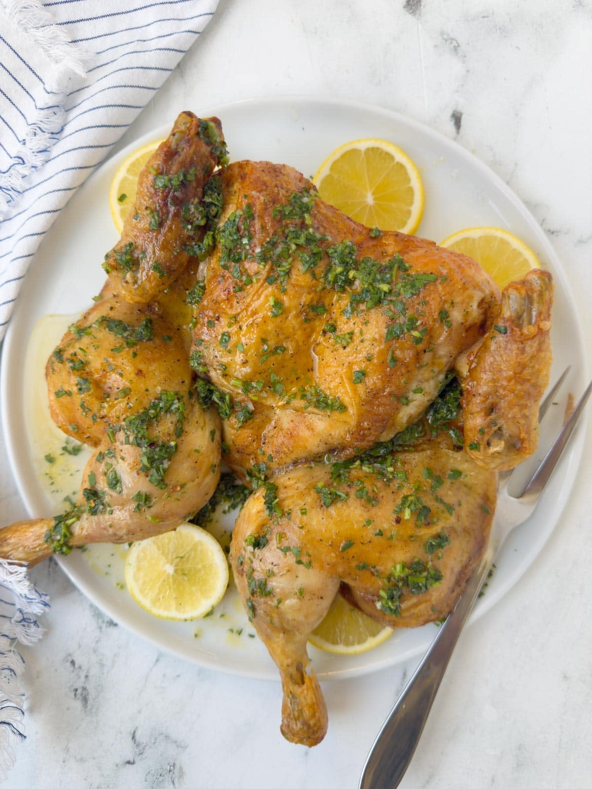 A golden spatchcock roasted chicken on a platter with lemon slices and green herbs,