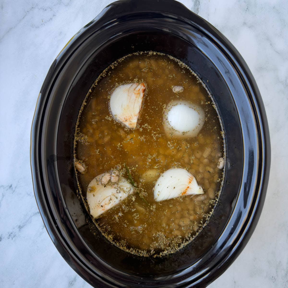 Broth and water added to a crock pot of dried beans. aromatics, and spices in a black ceramic insert.