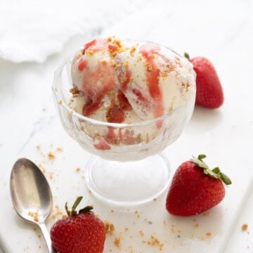 A glass bowl of strawberry cheesecake ice cream with strawberry sauce and buttered graham crumbles.