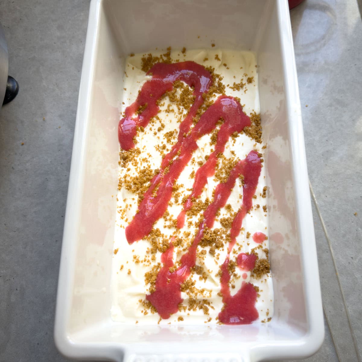 Layering ice cream, strawberry sauce, and graham crumbs into a loaf pan for making strawberry cheesecake ice cream.