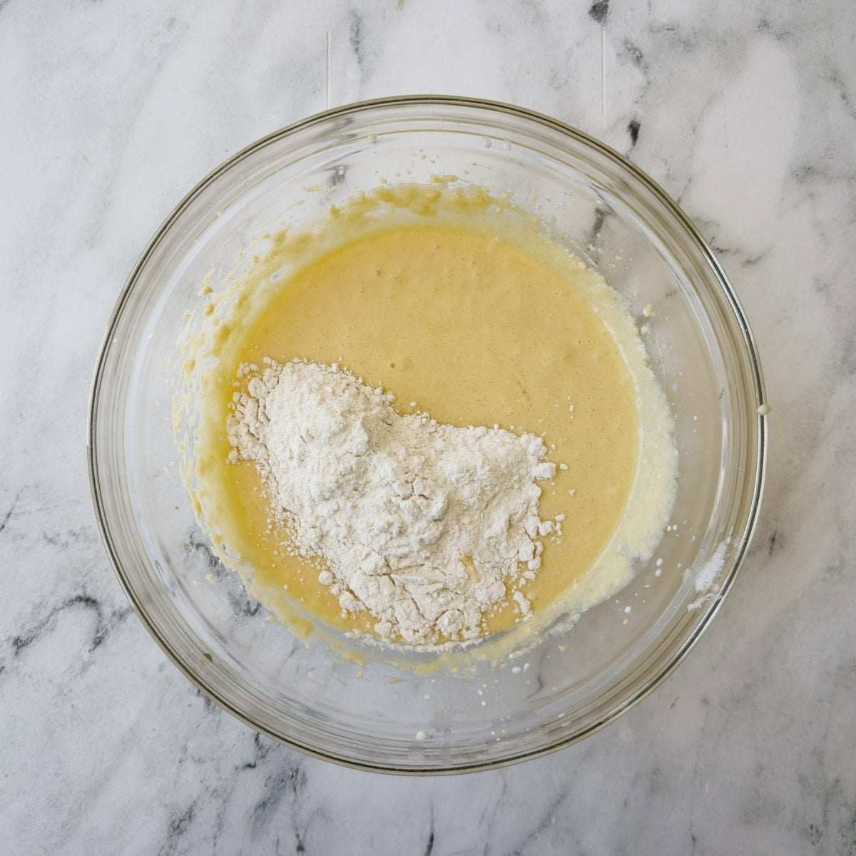 Golden cake batter in a glass bowl with flour being mixed in.